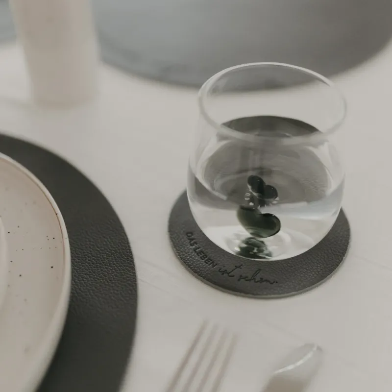 Glas auf dunkelgrauem Untersetzer mit Prägung ‚Das Leben ist schön‘ auf hell gedecktem Tisch.