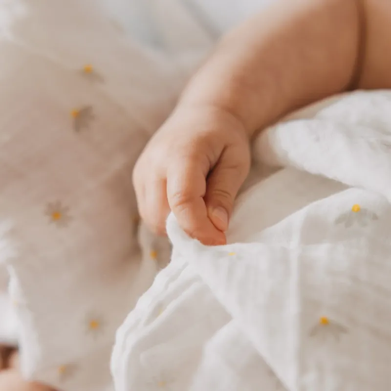 Kleine Babyhand greift nach einem weichen Musselintuch mit zartem Gänseblümchen-Print. Die Nahaufnahme hebt die feine Struktur und die angenehme Haptik des Stoffes hervor.